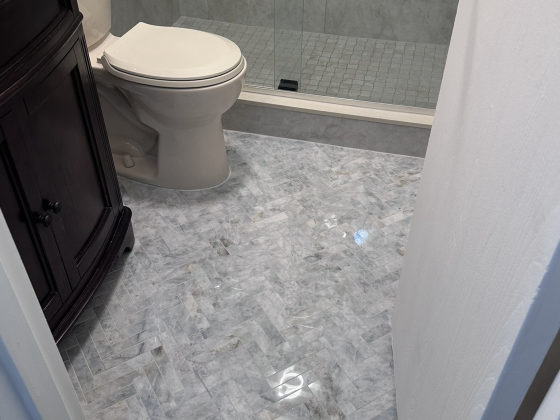 Modern bathroom with gray tile shower enclosure, white toilet, dark vanity with granite countertop, and marble-pattern floor tiles
